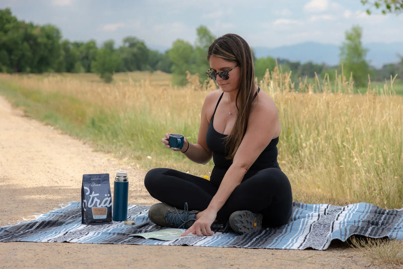 Woman sitting on a blanket outdoors with a coffee cup and products.